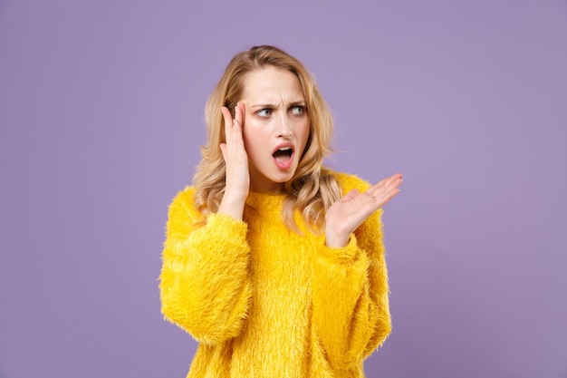 Mujer joven irritada disgustada con suéter de piel amarilla posando aislada en un retrato de estudio de fondo violeta pastel. Concepto de estilo de vida de las personas. Simulacros de espacio de copia. Difundir las manos mirando a un lado.