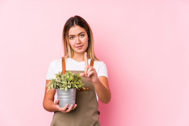 Mujer joven jardinero caucásico en una pared de color rosa que muestra el número uno con el dedo.