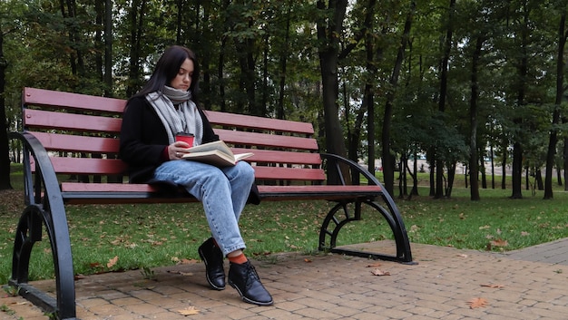 Mujer joven en jeans, abrigo y bufanda, en un banco del parque. Una mujer está leyendo un libro y tomando café u otra bebida caliente al aire libre sola.