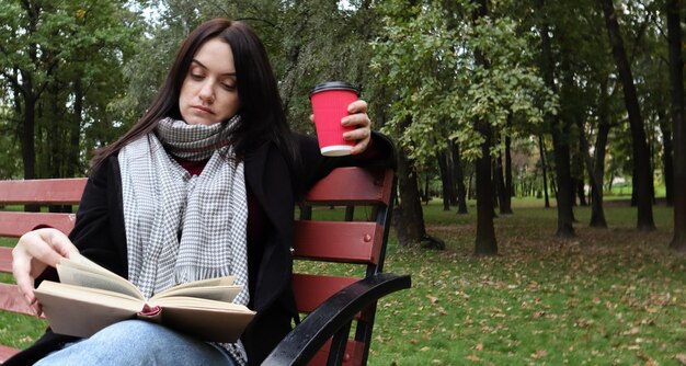 Mujer joven en jeans, abrigo y bufanda, en un banco del parque. Una mujer está leyendo un libro y tomando café u otra bebida caliente al aire libre sola.