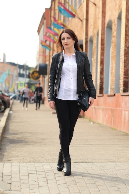 Mujer joven en jeans ajustados negros, camisa blanca y chaqueta de cuero camina por la calle