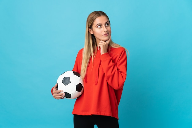 Mujer joven jugador de fútbol aislado en la pared azul mirando hacia arriba mientras sonríe