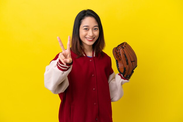 Mujer joven jugador vietnamita con guante de béisbol aislado sobre fondo amarillo sonriendo y mostrando el signo de la victoria