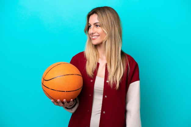 Mujer joven jugadora de baloncesto caucásica aislada de fondo azul mirando hacia un lado y sonriendo