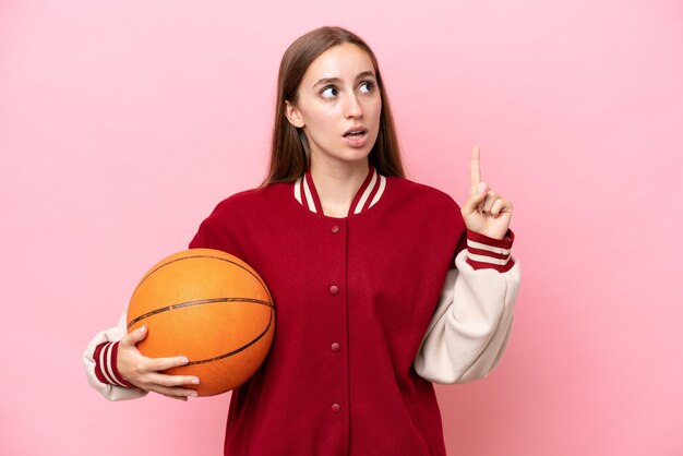 Mujer joven jugadora de baloncesto caucásica aislada en un fondo rosa pensando en una idea apuntando con el dedo hacia arriba