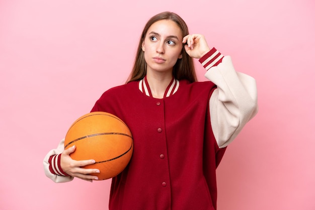 Mujer joven jugadora de baloncesto caucásica aislada en un fondo rosa que tiene dudas y piensa