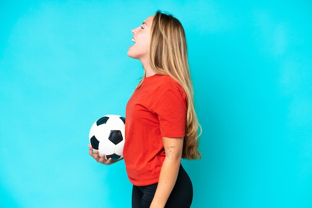 Mujer joven jugadora de fútbol aislada de fondo azul riéndose en posición lateral