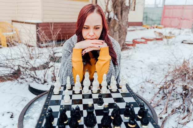 Mujer joven jugando al ajedrez en el patio Hembra envuelta en cuadros grises sentada en la calle jugando en el juego de mesa en la temporada de invierno