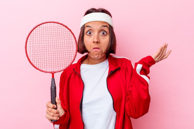 Mujer joven jugando al bádminton aislado en la pared rosada se encoge de hombros y abre los ojos confundidos