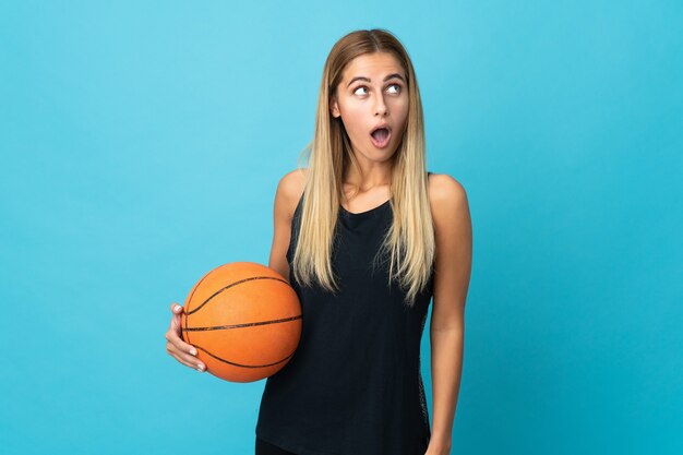 Mujer joven jugando baloncesto aislado en la pared blanca mirando hacia arriba y con expresión de sorpresa