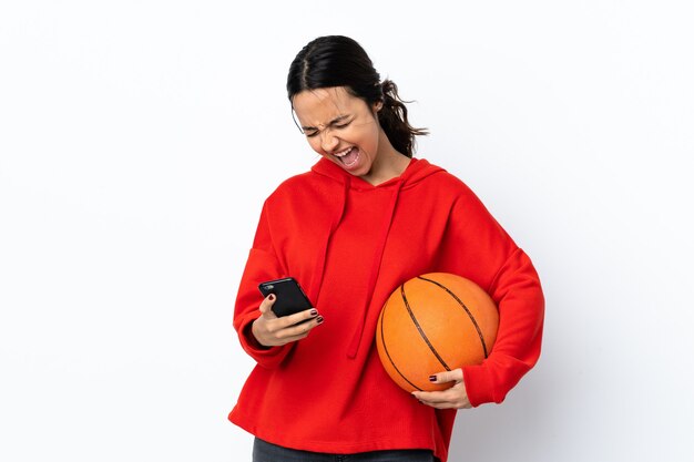 Mujer joven jugando baloncesto sobre blanco aislado con teléfono en posición de victoria