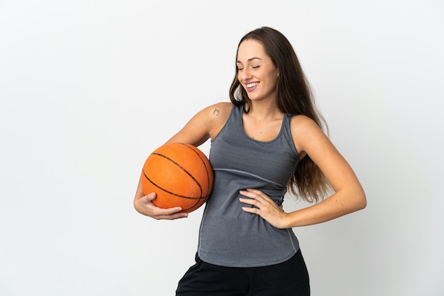 Mujer joven jugando baloncesto sobre fondo blanco aislado posando con los brazos en la cadera y sonriendo