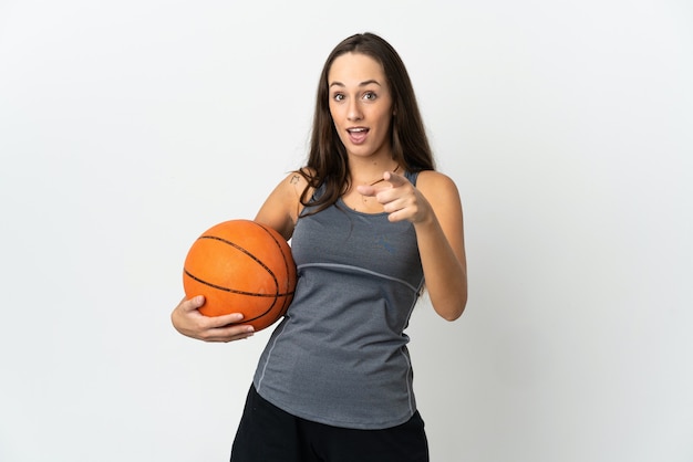 Mujer joven jugando baloncesto sobre fondo blanco aislado sorprendido y apuntando hacia el frente