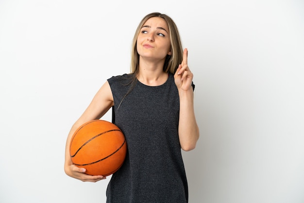 Mujer joven jugando baloncesto sobre pared blanca aislada con los dedos cruzando y deseando lo mejor