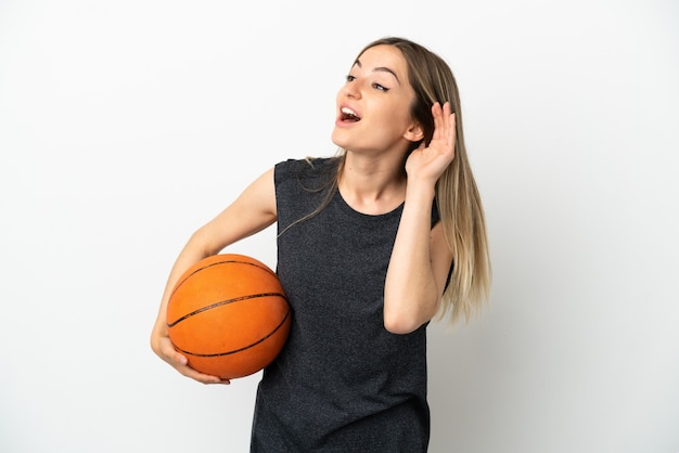 Mujer joven jugando baloncesto sobre pared blanca aislada escuchando algo poniendo la mano en la oreja