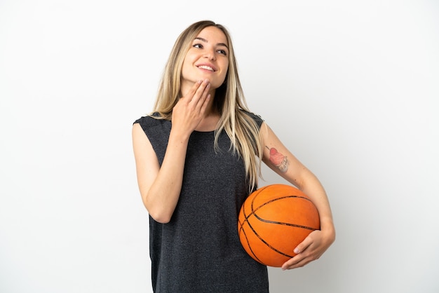 Mujer joven jugando baloncesto sobre pared blanca aislada mirando hacia arriba mientras sonríe