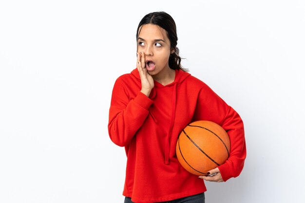 Mujer joven jugando baloncesto sobre pared blanca aislada susurrando algo con gesto de sorpresa mientras mira hacia el lado