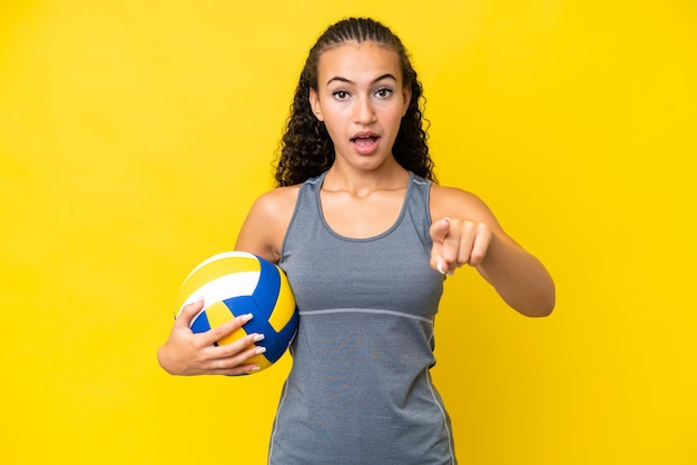 Mujer joven jugando voleibol aislado sobre fondo amarillo sorprendido y apuntando al frente