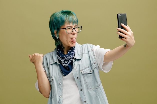 Mujer joven juguetona con gafas bandana en el cuello manteniendo el puño en el aire estirando el teléfono móvil mostrando la lengua tomando selfie aislado sobre fondo verde oliva