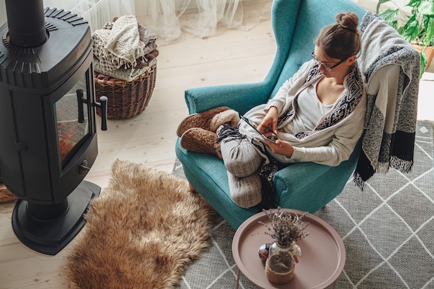 Mujer joven junto a la chimenea, sentada en un cómodo sillón, con una manta caliente, usando una tableta