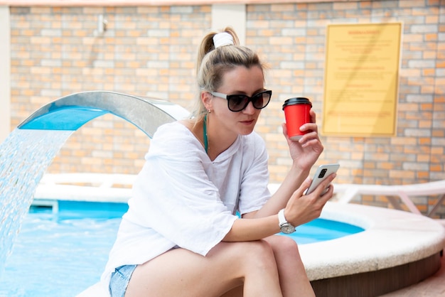 Una mujer joven junto a la piscina con un aparato y café en las manos. Está de vacaciones.