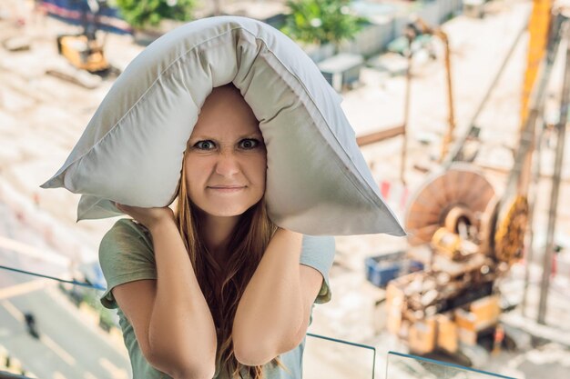 Una mujer joven junto a la ventana molesta por los trabajos de construcción afuera me tapó los oídos con una almohada. Concepto de ruido