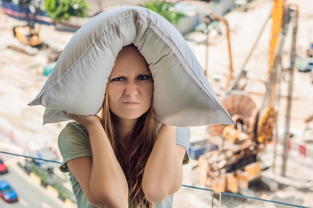 Una mujer joven junto a la ventana molesta por los trabajos de construcción afuera me tapó los oídos con una almohada. Concepto de ruido