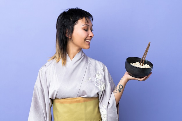 Mujer joven con kimono sobre azul aislado con expresión feliz mientras sostiene un tazón de fideos con palillos