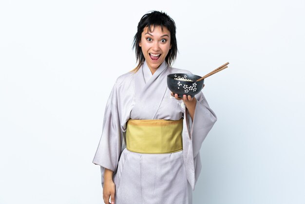 Mujer joven con kimono sosteniendo un tazón de fideos sobre blanco aislado con sorpresa y expresión facial conmocionada