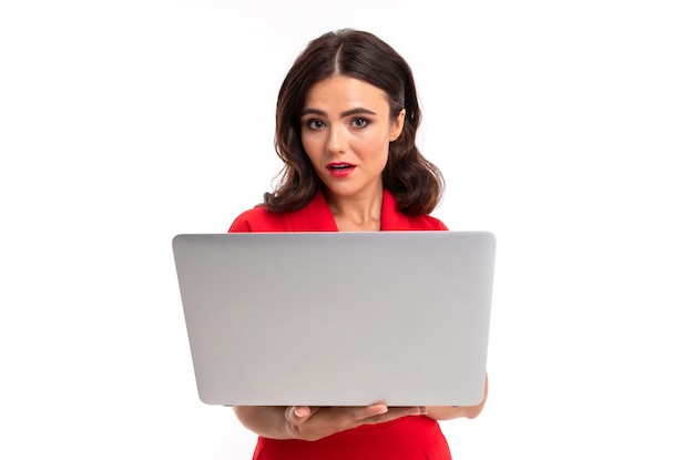 Foto una mujer joven con labios rojos, maquillaje brillante, cabello largo ondulado oscuro, con un traje rojo, se para con una laptop blanca