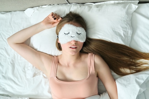 Mujer joven con largo cabello rubio con antifaz para dormir en sus ojos durmiendo en la cama blanca, vista superior