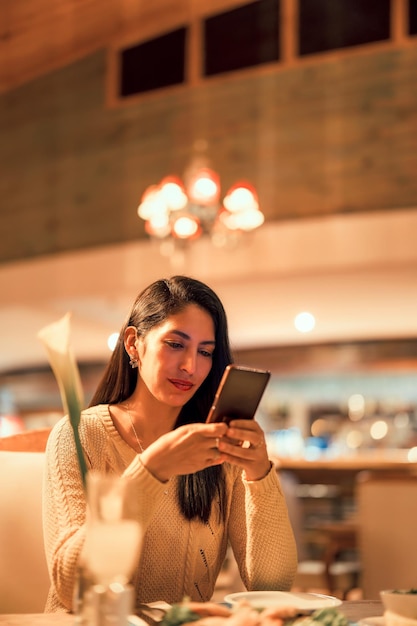Mujer joven latina sentada en un café o restaurante usando un retrato vertical de teléfono inteligente
