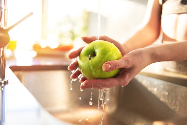 Mujer joven se lava, con agua corriente, una manzana en el fregadero de la cocina iluminado por el sol
