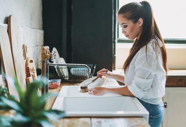 Mujer joven lava platos con cepillo de madera con cerdas naturales en la ventana de la cocina. concepto de cero residuos