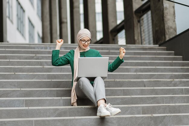 Mujer joven levanta la mano de buenas noticias mientras trabaja en una computadora portátil al aire libre