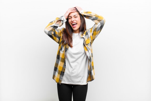 Mujer joven levantando las manos a la cabeza, con la boca abierta, sintiéndose extremadamente afortunado, sorprendido, emocionado y feliz aislado en la pared blanca