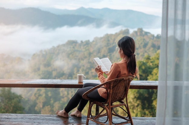 Mujer joven leyendo un libro cerca de la ventana y mirando la vista de la montaña en la casa de campo en el amanecer de la mañana. SoloTravel, viaje, viaje y concepto relajante