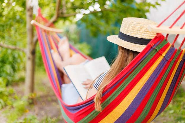 Mujer joven leyendo un libro mientras se acuesta en una cómoda hamaca en el jardín verde