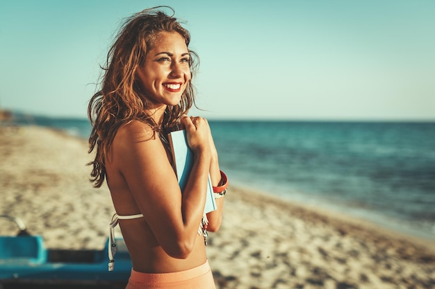 Mujer joven leyendo un libro en la playa de arena y pensando con una sonrisa en su rostro.