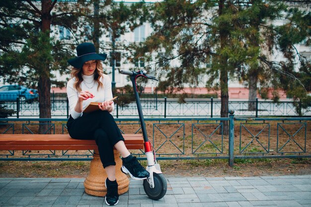 Mujer joven leyendo un libro y sentada en un banco con scooter eléctrico en la ciudad