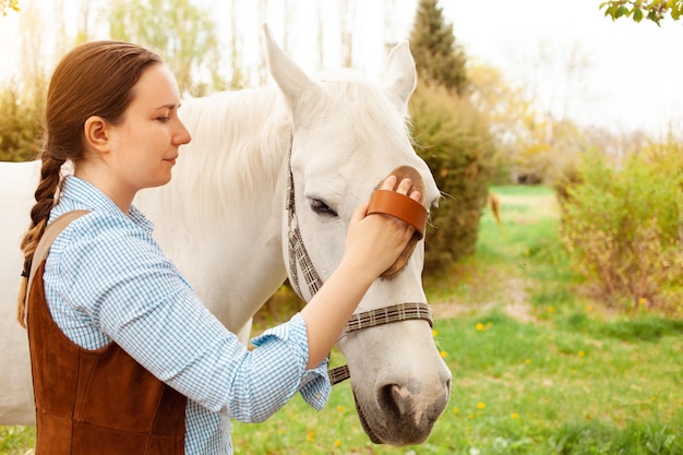 Una mujer joven limpia un caballo blanco con un cepillo amarillo en la naturaleza