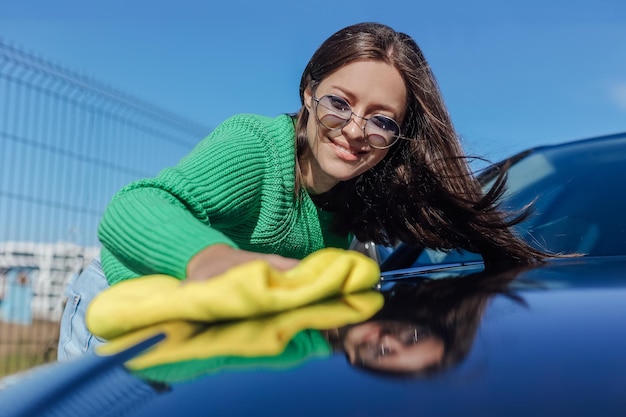 Una mujer joven limpiando y puliendo el capó de su auto con un concepto de tela de microfibra de cuidado del auto