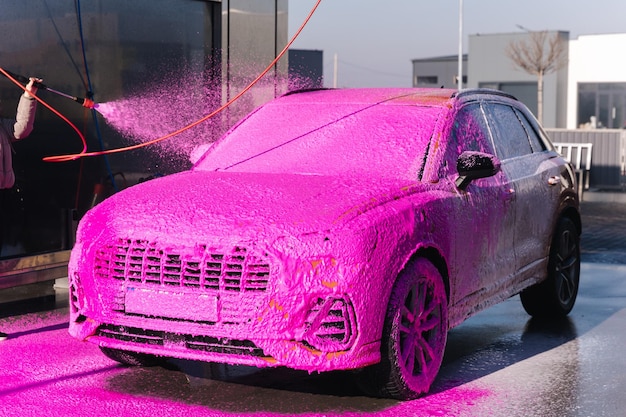Mujer joven limpiando su auto en autoservicio de lavado de autos jabón esparcido femenino usando espuma rosa con rociador de chorro