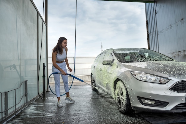 Mujer joven limpiando su coche con una manguera con spray de espuma y agua a presión en el lavado manual de coches de la suciedad