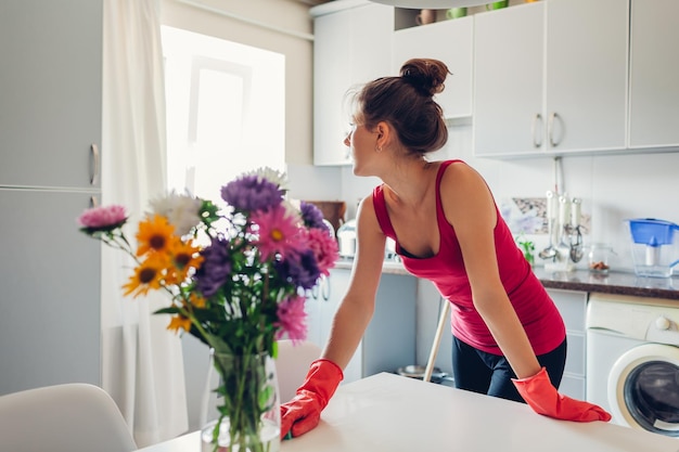 Mujer joven limpiando la superficie de la mesa con esponja en la cocina moderna decorada con flores.