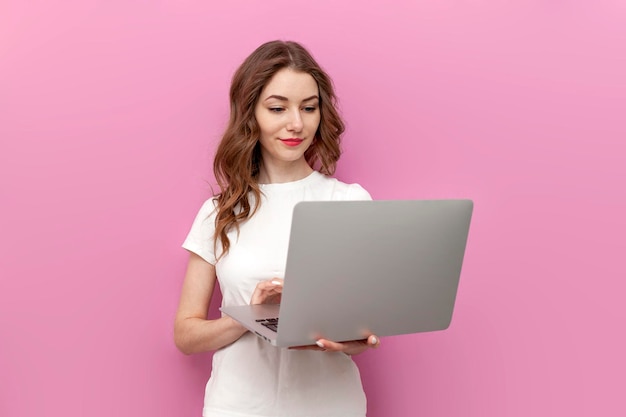 Una mujer joven y linda con una camiseta blanca usa una computadora portátil sobre un fondo rosa aislado y tipos de profesionales independientes