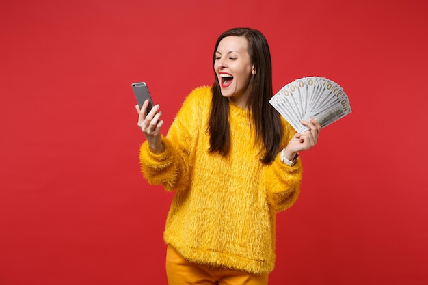 Mujer joven llena de alegría en suéter de piel mirando en teléfono móvil mantenga ventilador de dinero en billetes de dólar dinero en efectivo aislado sobre fondo rojo. Concepto de estilo de vida de emociones sinceras de personas. Simulacros de espacio de copia.