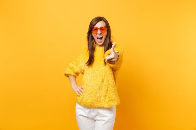 Mujer joven llena de alegría en suéter de piel, pantalón blanco, gafas de corazón naranja gritando, mostrando el pulgar hacia arriba aislado sobre fondo amarillo brillante. Personas sinceras emociones, concepto de estilo de vida. Área de publicidad.