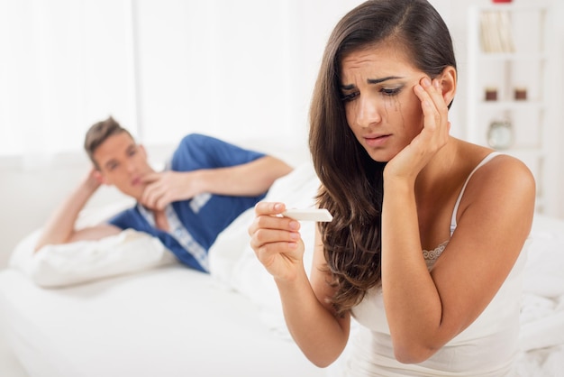 Mujer joven llorando sentada en la cama y mirando una prueba de embarazo mientras que en el fondo ve a su pareja acostada en la cama y mirando a la mujer.