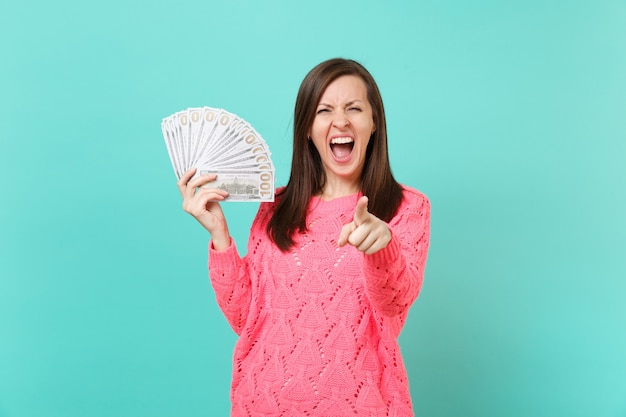 Mujer joven loca en suéter rosa gritando, apuntando con el dedo índice a la cámara mantenga un montón de billetes de dólares, dinero en efectivo aislado sobre fondo azul. Concepto de estilo de vida de personas. Simulacros de espacio de copia.